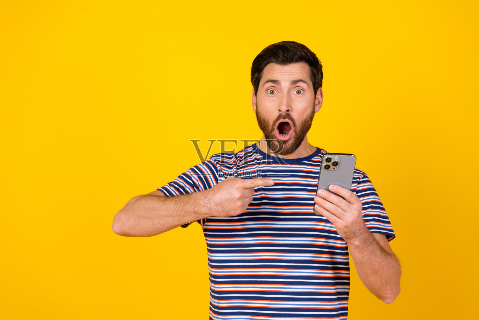 一个穿着条纹t恤的欣喜若狂的家伙的肖像，在鲜艳的黄色背景下，他盯着智能手机看照片摄影图片