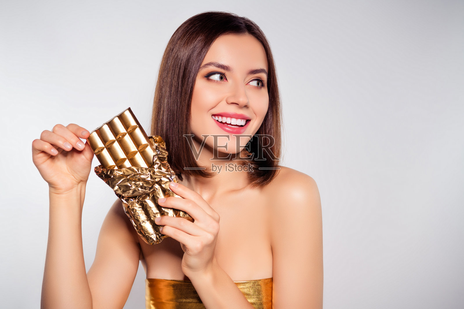 照片中快乐的女孩牙齿微笑着拿着金色的巧克力棒，看起来空无一物，孤立的灰色背景照片摄影图片