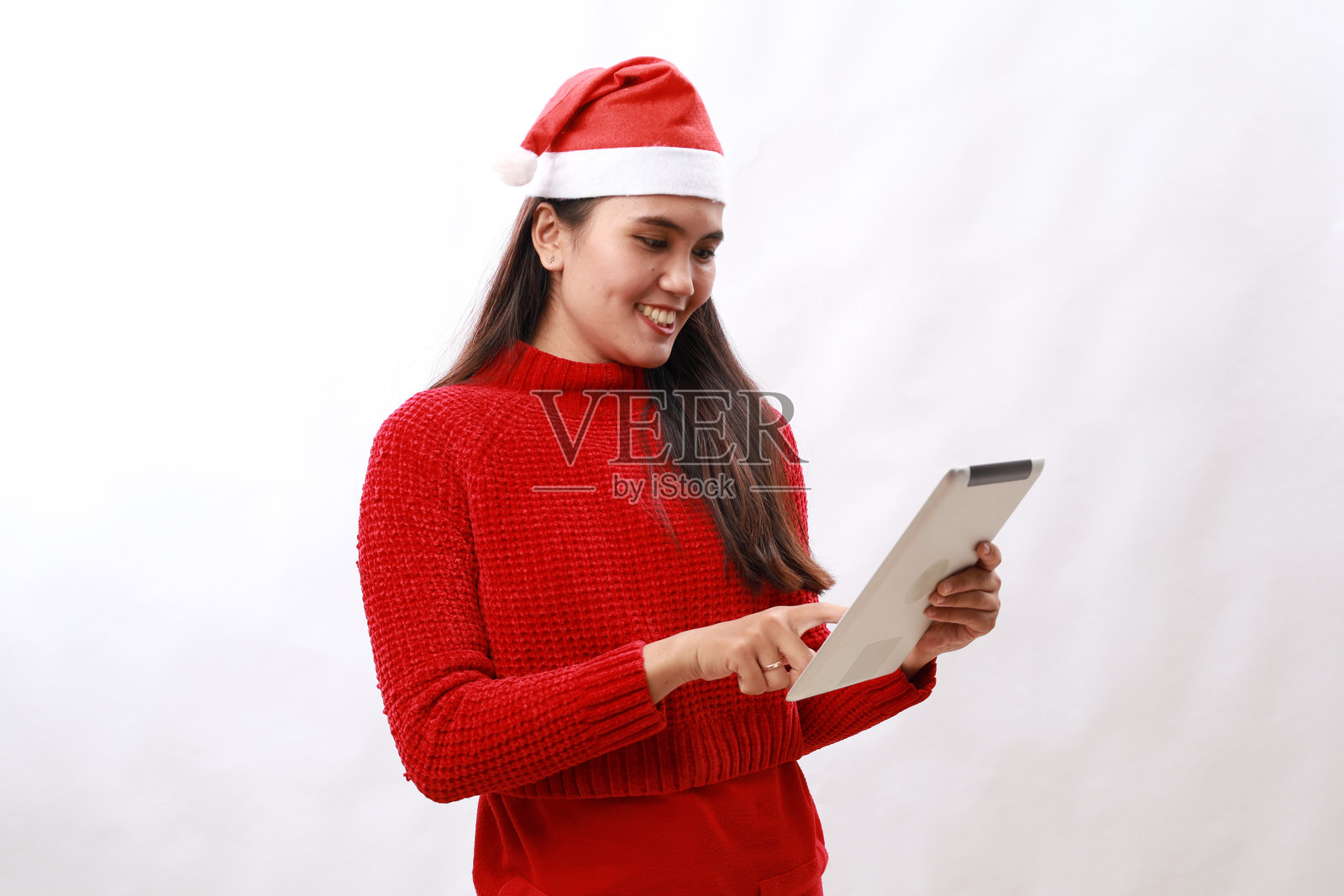 年轻的亚洲女子穿着红色高领毛衣，戴着圣诞老人帽，用一台孤立的白色背景的平板电脑照片摄影图片