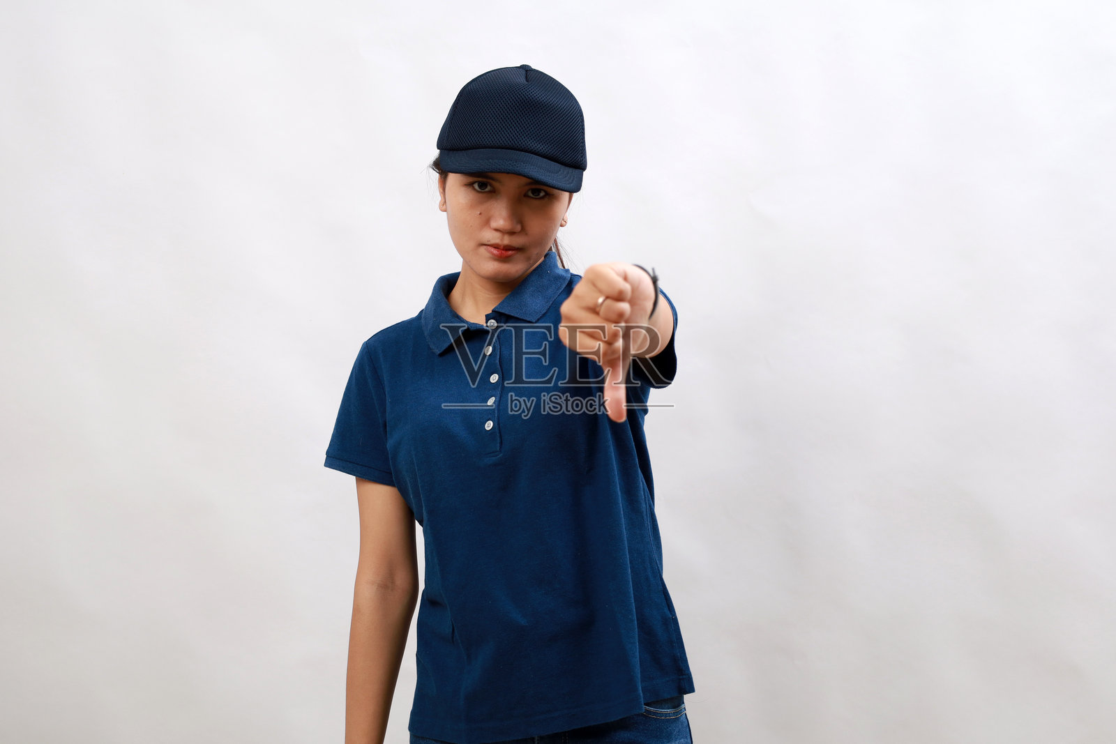 年轻的亚洲女快递员穿着快递制服，戴着帽子，看起来不高兴和生气，用拇指向下的手势表示拒绝和否定。糟糕的表达。照片摄影图片