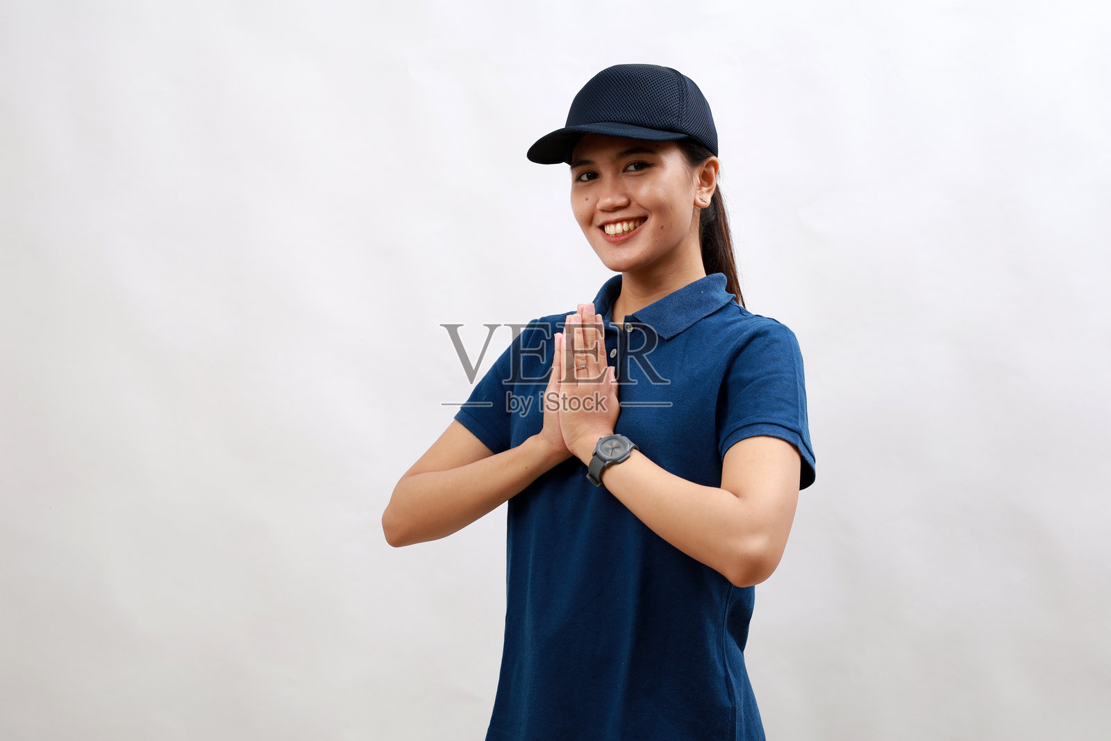 年轻的亚洲女快递员穿着快递制服，戴着帽子，双手合拢祈祷，请求原谅，微笑着，自信地笑着。照片摄影图片