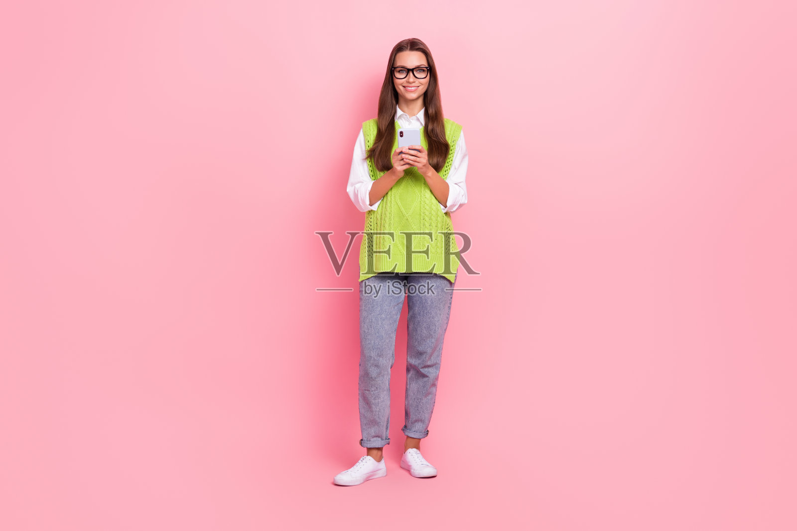一个开朗的女人，留着长发，穿着绿色马甲牛仔裤，拿着智能手机，孤立在粉红色的背景上照片摄影图片