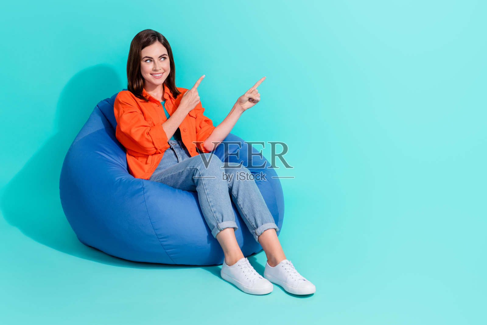 全身照片梦幻闪亮的女士穿着橙色衬衫坐在豆袋指向空空间孤立的蓝绿色背景照片摄影图片