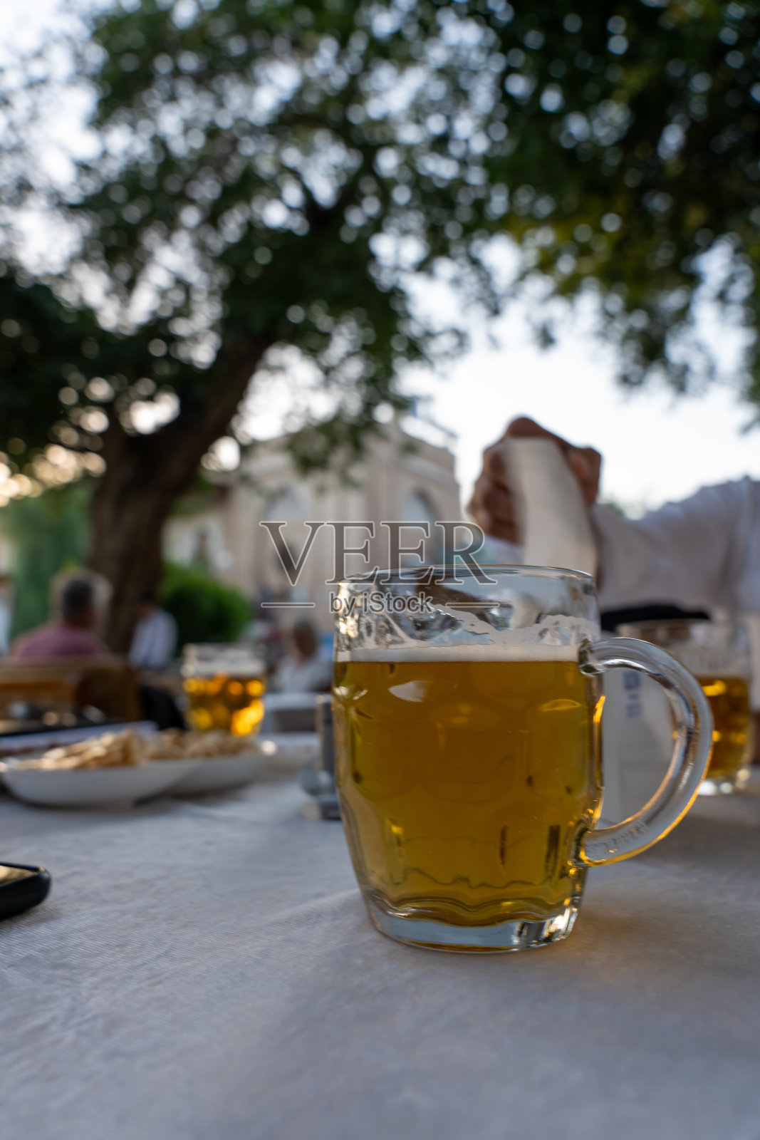 一棵树前的桌子上放着一杯啤酒。现场是随意和放松的，人们围坐在桌子周围，享受着他们的饮料照片摄影图片