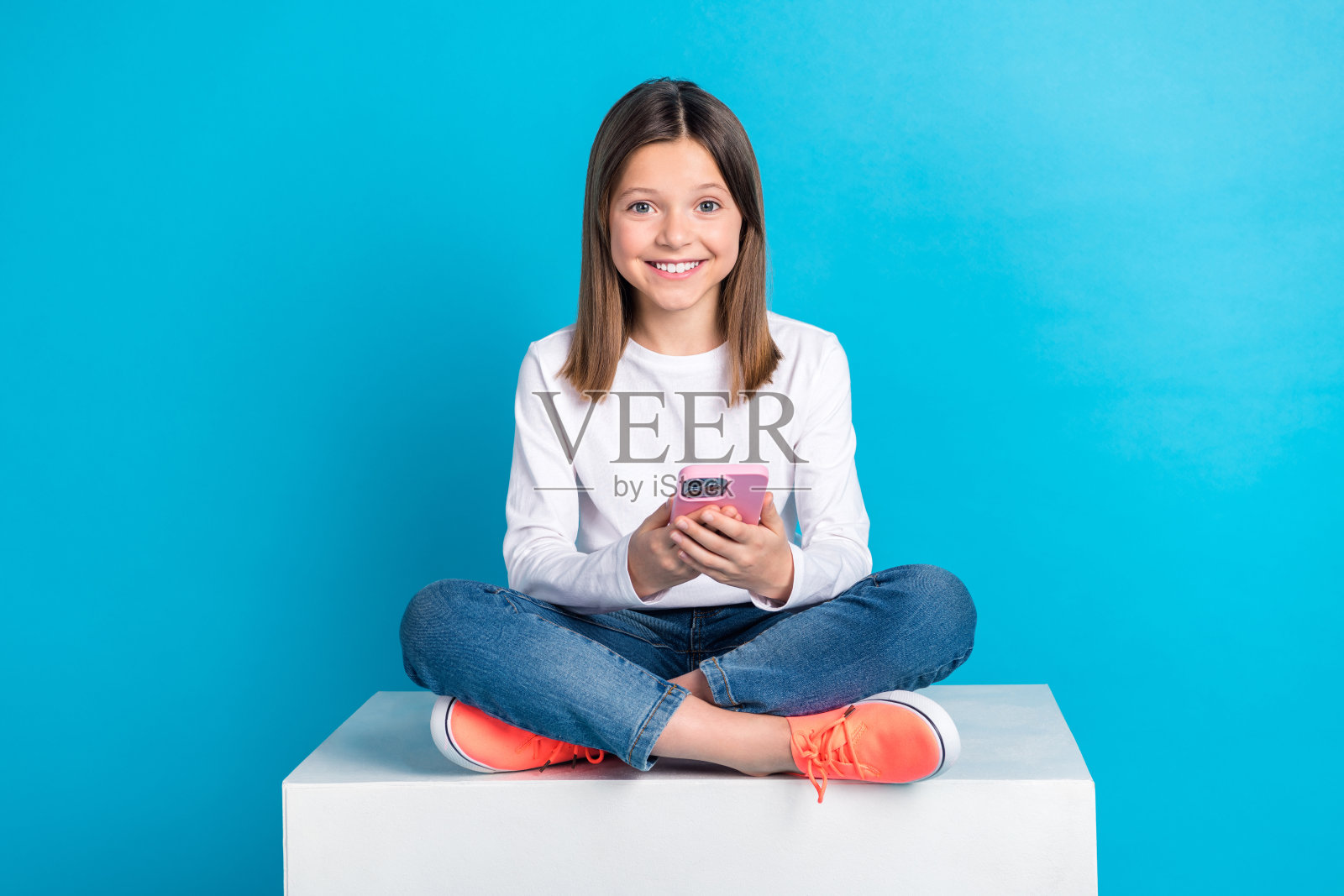 快乐的女孩穿白色长袖牛仔裤的全尺寸照片，阅读通知，坐在立方体上孤立的蓝色背景照片摄影图片