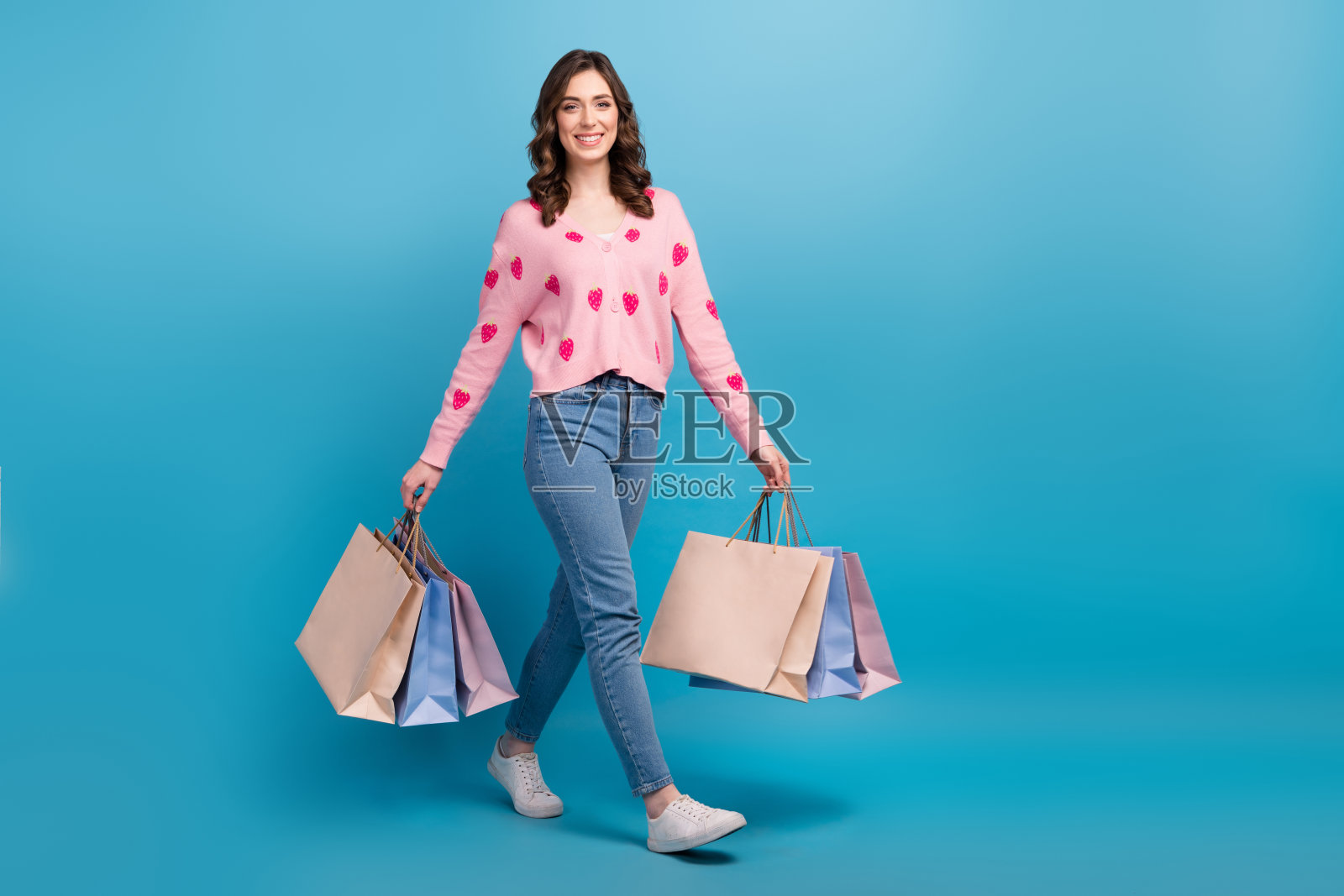 全身照片可爱甜美的女人穿着粉红色的开襟羊毛衫抱着购物者走空旷的空间孤立的蓝色背景照片摄影图片