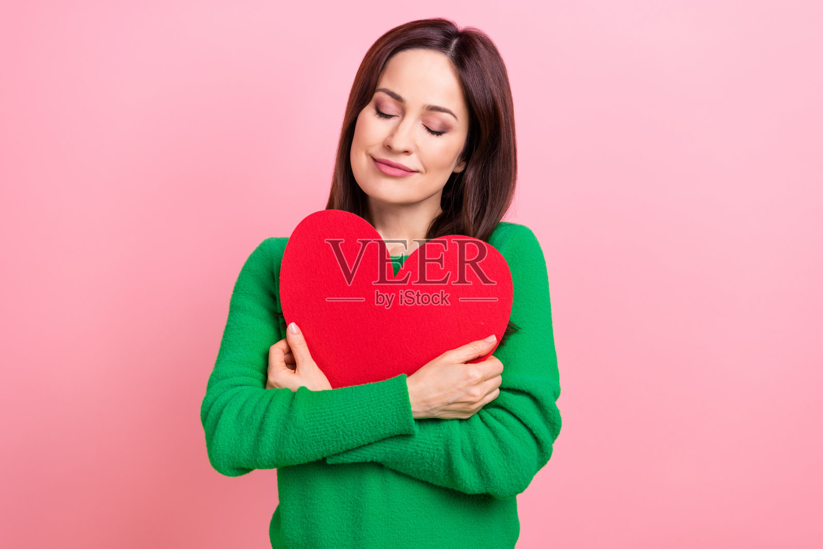 可爱可爱的女士穿着绿色套衫拥抱红色的心，闭上眼睛孤立的粉红色背景的照片照片摄影图片