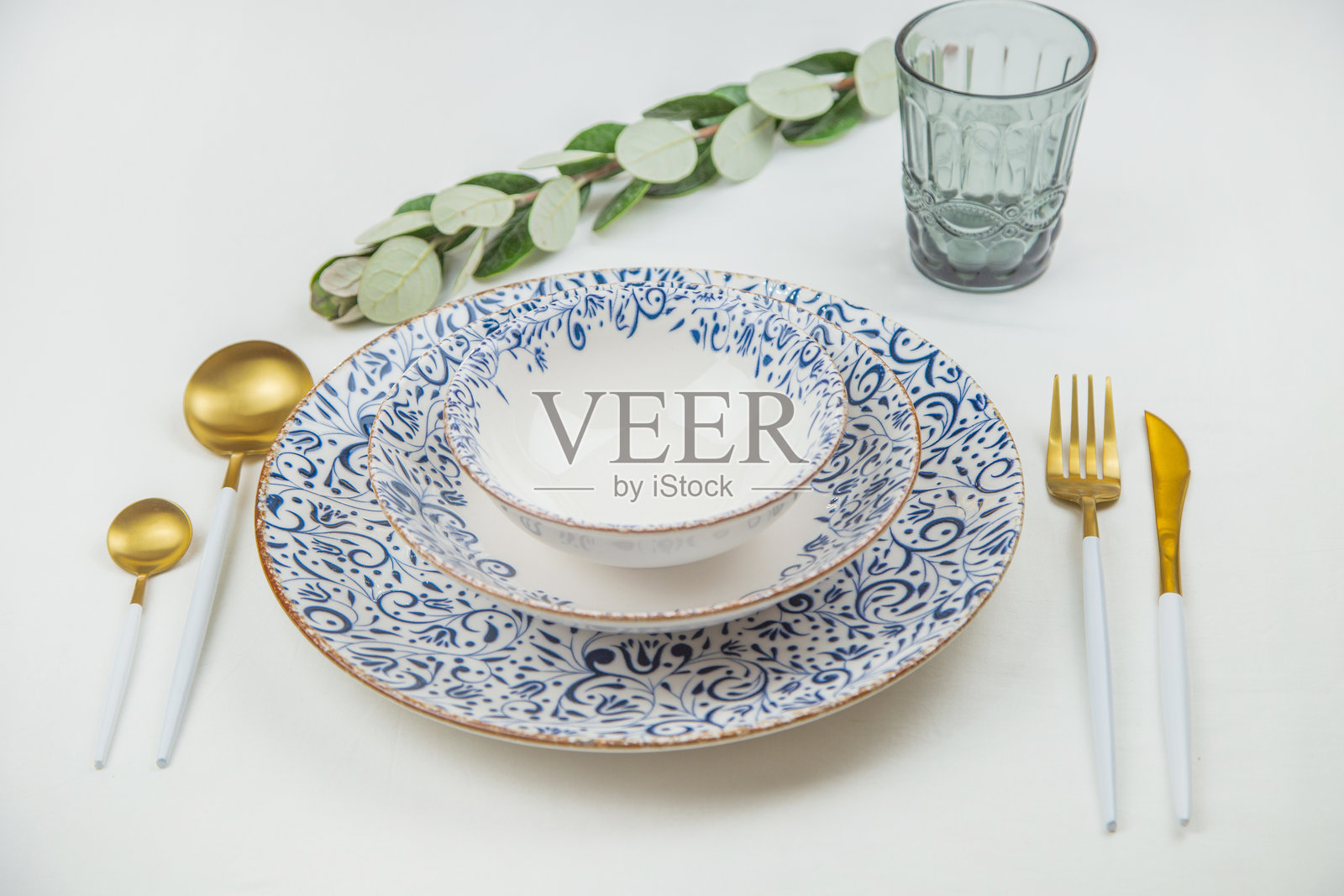 一套边缘有蓝色图案的白色盘子互相叠在一起，勺子、刀子和叉子放在两边，它们后面是一个蓝色的玻璃杯和白色背景上的绿色嫩芽。照片摄影图片