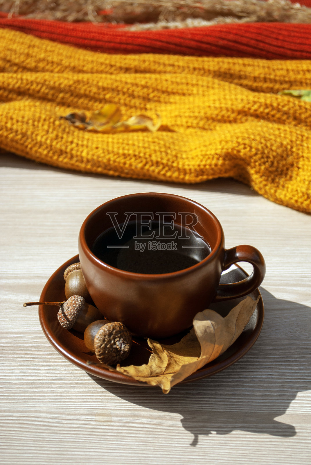 棕色的杯子上放着一碟热咖啡，枯萎的橡子，橙色和红色的针织毛衣放在浅色的木质表面上照片摄影图片