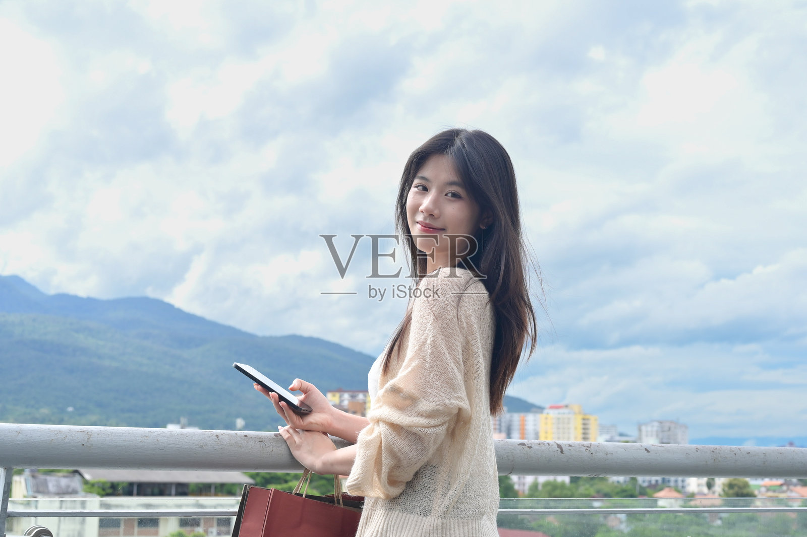 年轻女子手持手机和购物袋站在户外屋顶露台上的肖像，背景是城市景观照片摄影图片