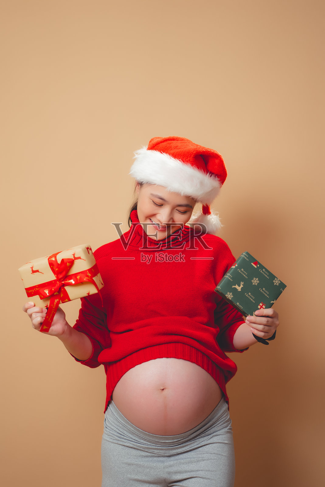 亚洲孕妇戴着圣诞老人的帽子，开心地微笑着，拿着礼盒，露出肚子，孤立的米色背景。照片摄影图片
