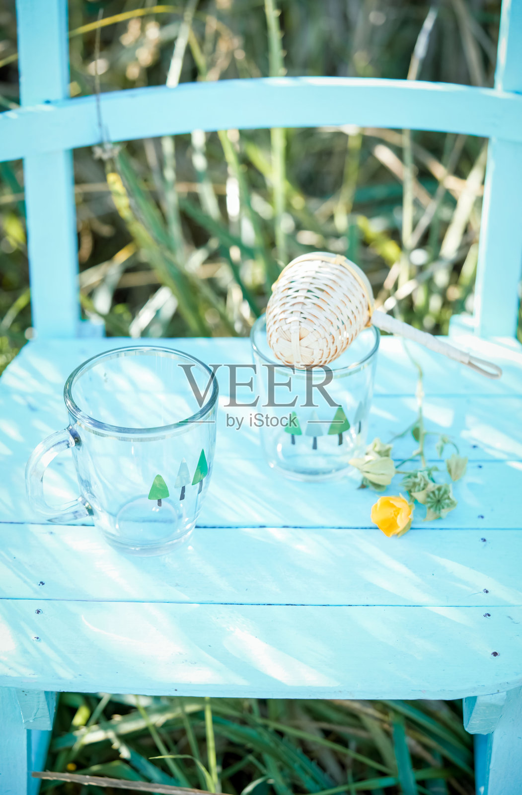 透明的马克杯，上面画着松树、柳条茶过滤器、鲜花，放在田野里的浅蓝色椅子上。照片摄影图片