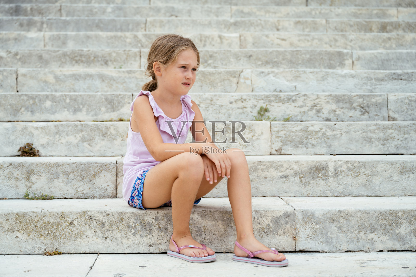 一个小女孩坐在石阶上，穿着浅紫色上衣和粉色人字拖，显得若有所思。照片摄影图片