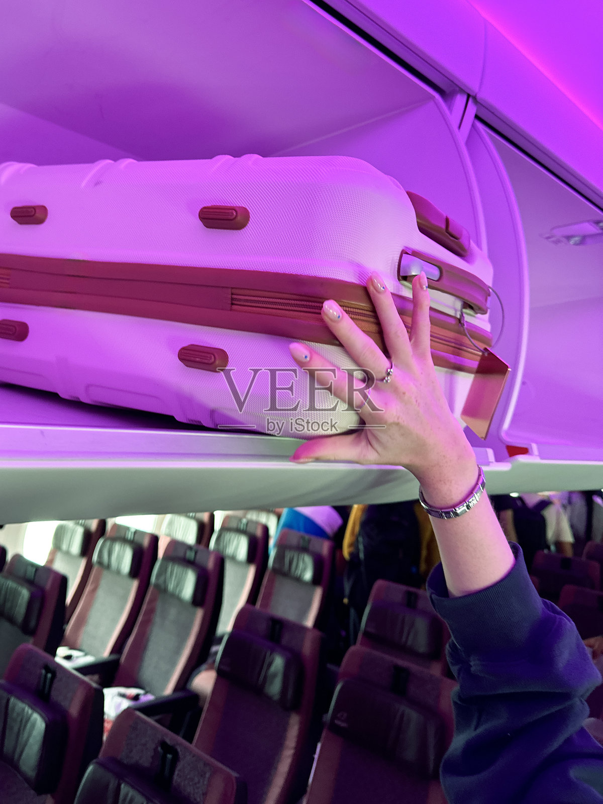 特写照片中，一个无法辨认的女人把带轮子的行李箱放在飞机机舱上方的行李舱里，粉红色的霓虹灯照亮了舒缓的登机心情，一排座位，聚焦在前景上照片摄影图片