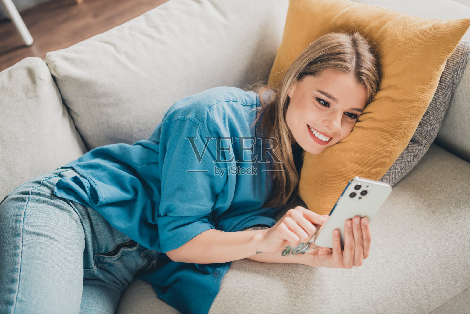 美丽快乐的金发女人在家里放松，躺在舒适柔软的沙发和枕头上，在客厅里看tiktoks照片摄影图片