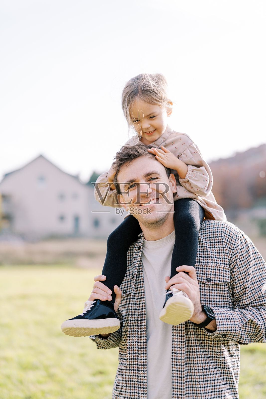 在阳光明媚的草地上，微笑的小女孩抚摸着爸爸的头发，爸爸把她扛在肩上照片摄影图片
