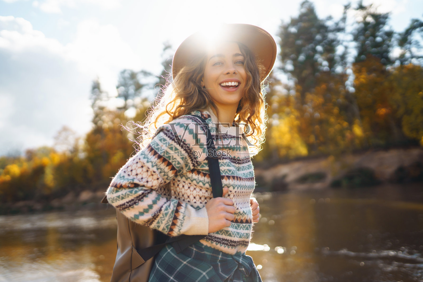 一个年轻的女旅行者在河边享受着一个阳光明媚的秋日下午，背着背包，脸上带着欢快的微笑。照片摄影图片