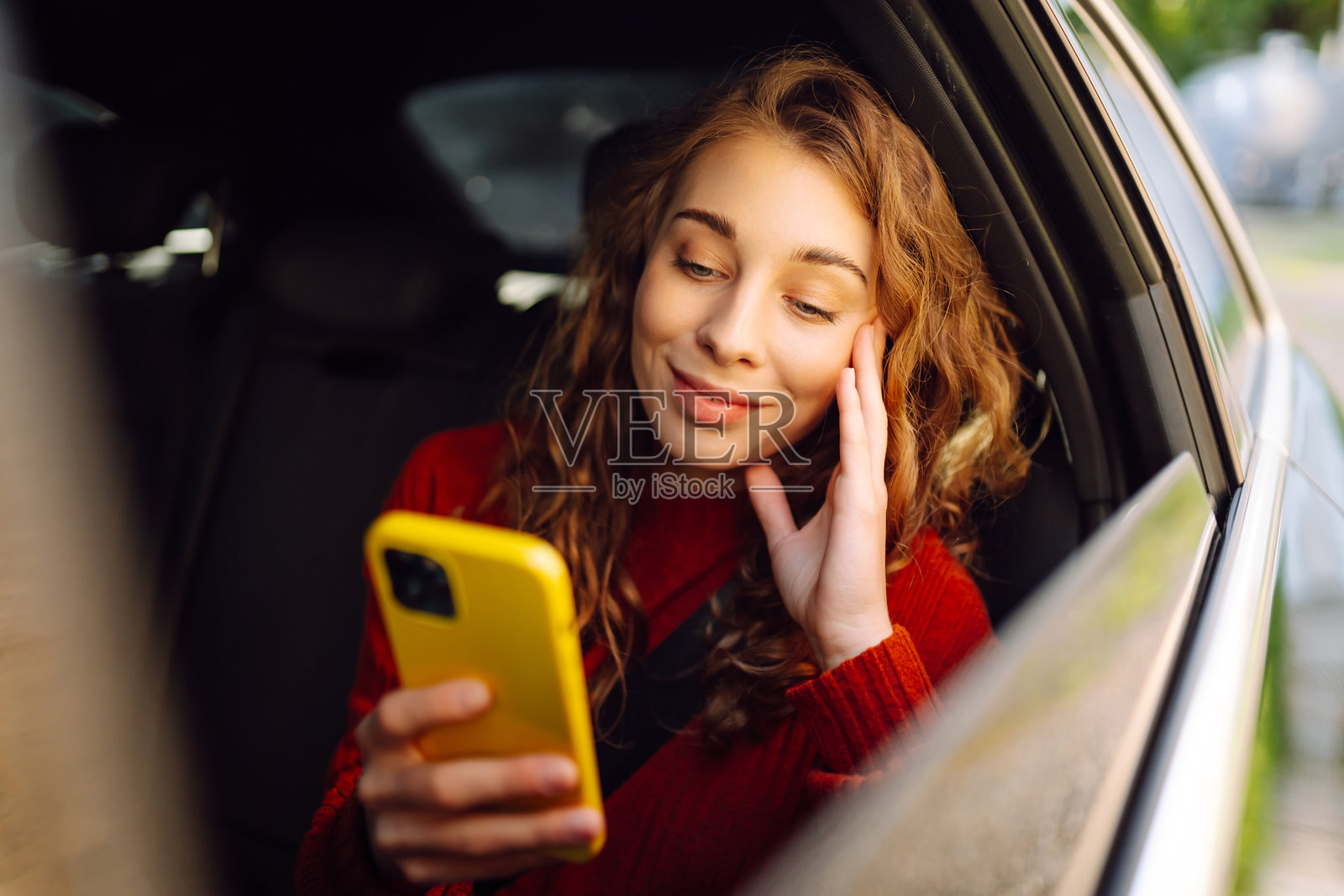 一位卷发的年轻女子坐在车里玩智能手机，享受着阳光明媚的一天。照片摄影图片