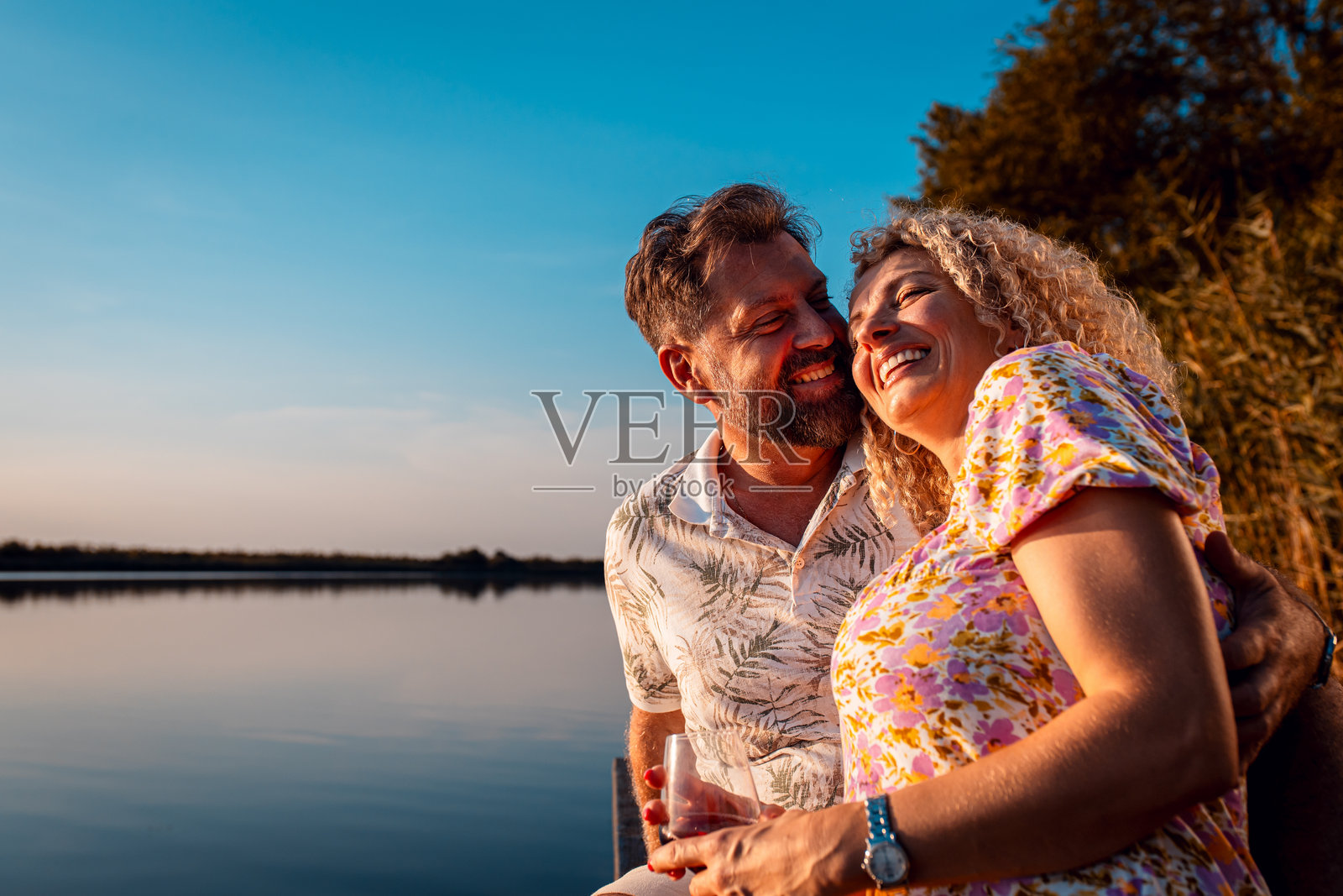 一对微笑的成年夫妇在日落时分坐在湖边的码头上喝酒。照片摄影图片