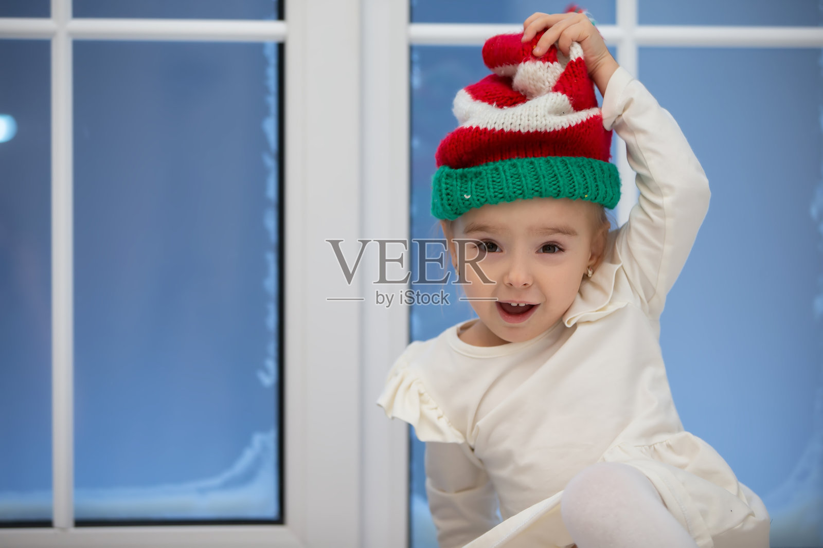 一个有趣的小女孩戴着圣诞帽，背景是一扇下雪的窗户。照片摄影图片