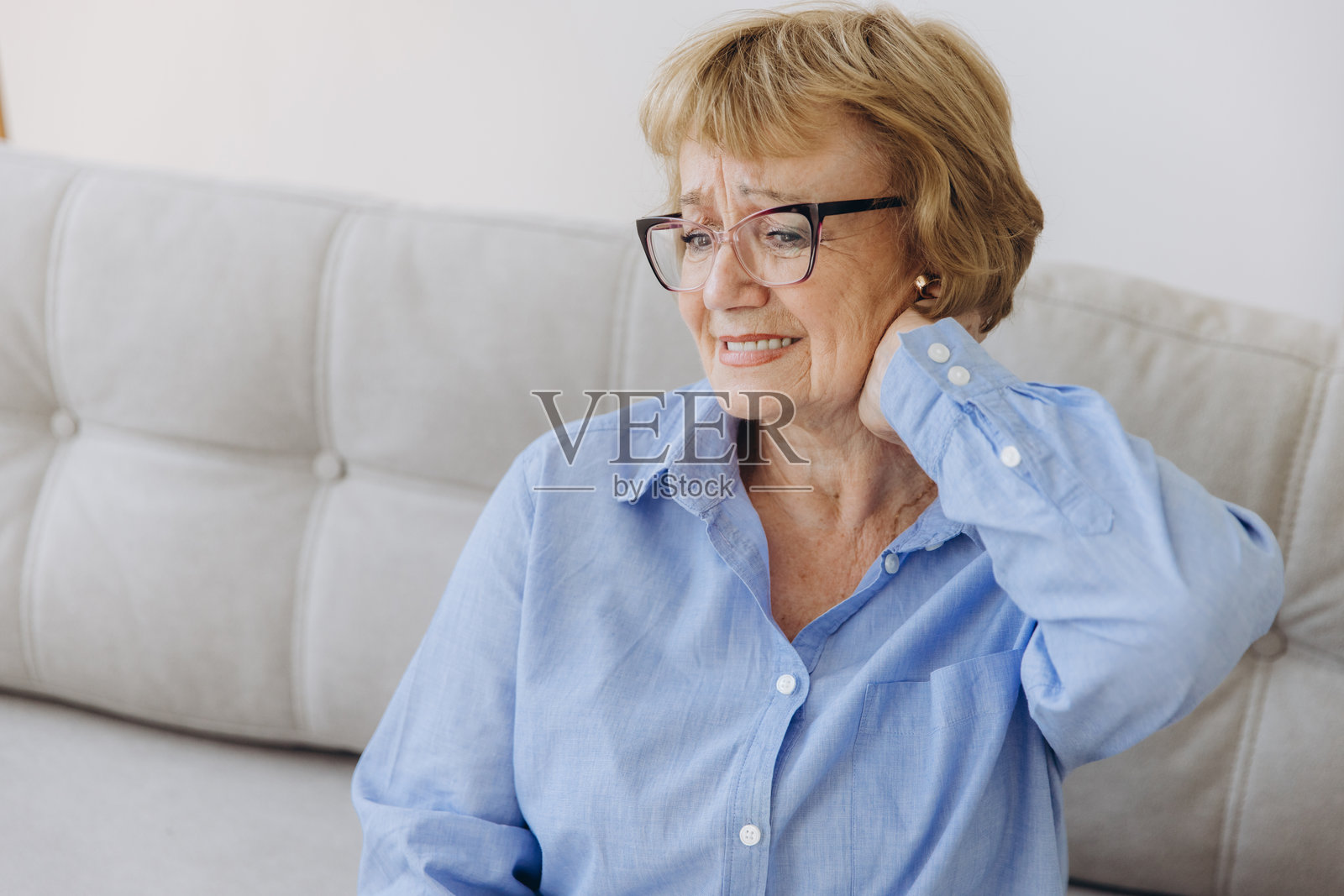 患病的老年成年妇女坐在沙发上摸脖子有肩颈疼痛。保健和医药理念照片摄影图片