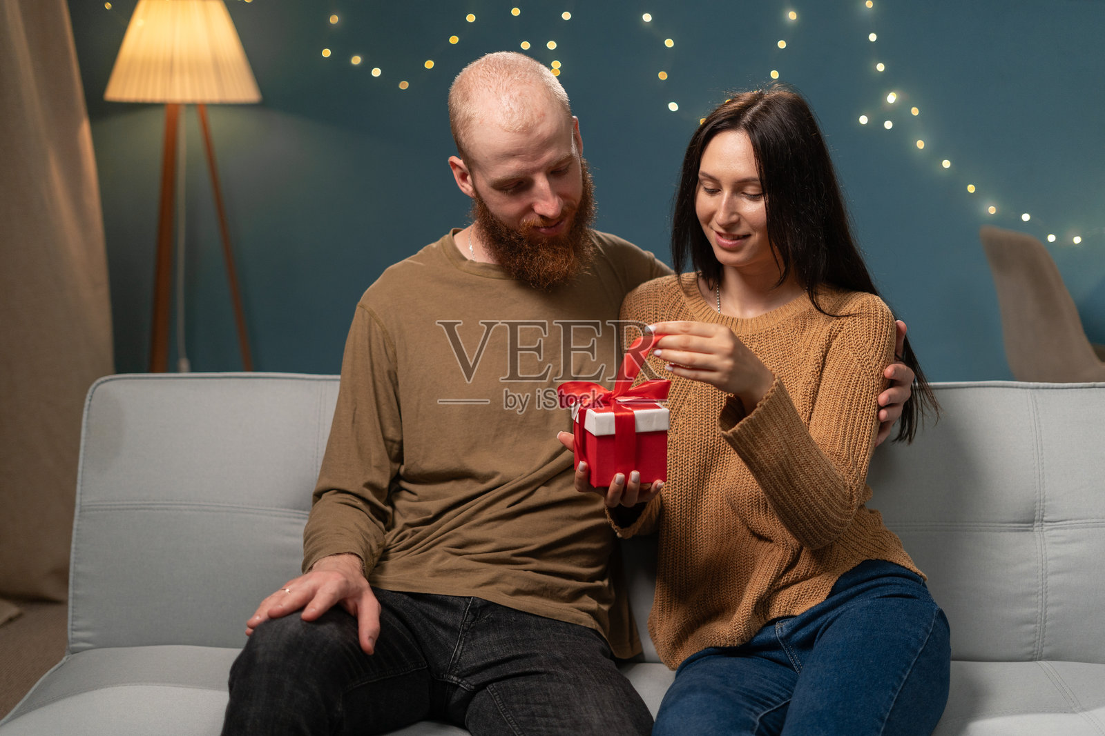 年轻女子和丈夫一起打开礼盒，坐在家里的沙发上送生日礼物。晚上在家里相爱的情侣照片摄影图片