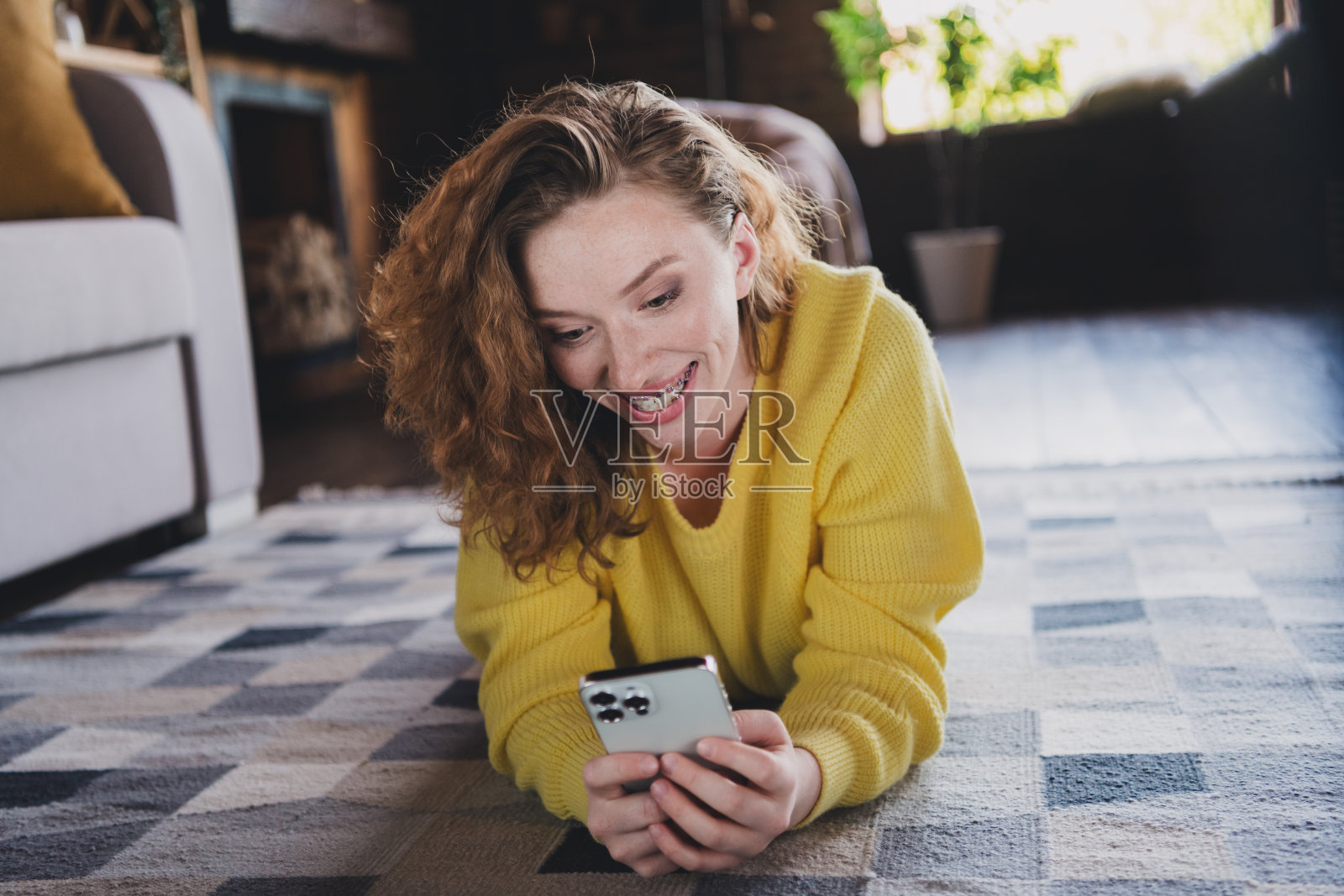 迷人的年轻女性躺在地板上的全尺寸照片，手持装置穿着黄色毛衣舒适的日光家庭室内客厅照片摄影图片
