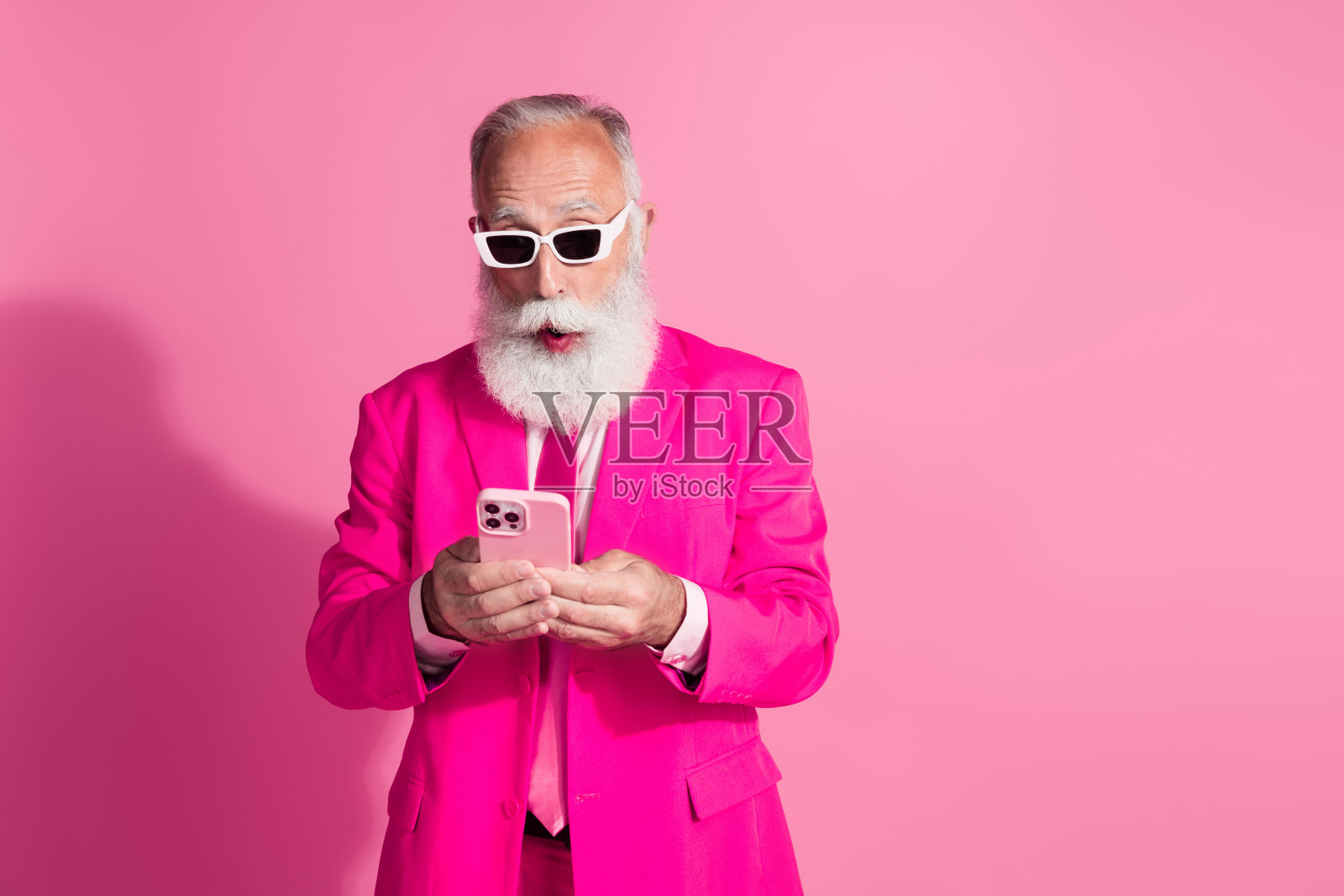 照片中严肃的老酷男人绅士男友惊讶地拿着装置戴着太阳镜时髦的粉色西装孤立在柔和的颜色背景照片摄影图片