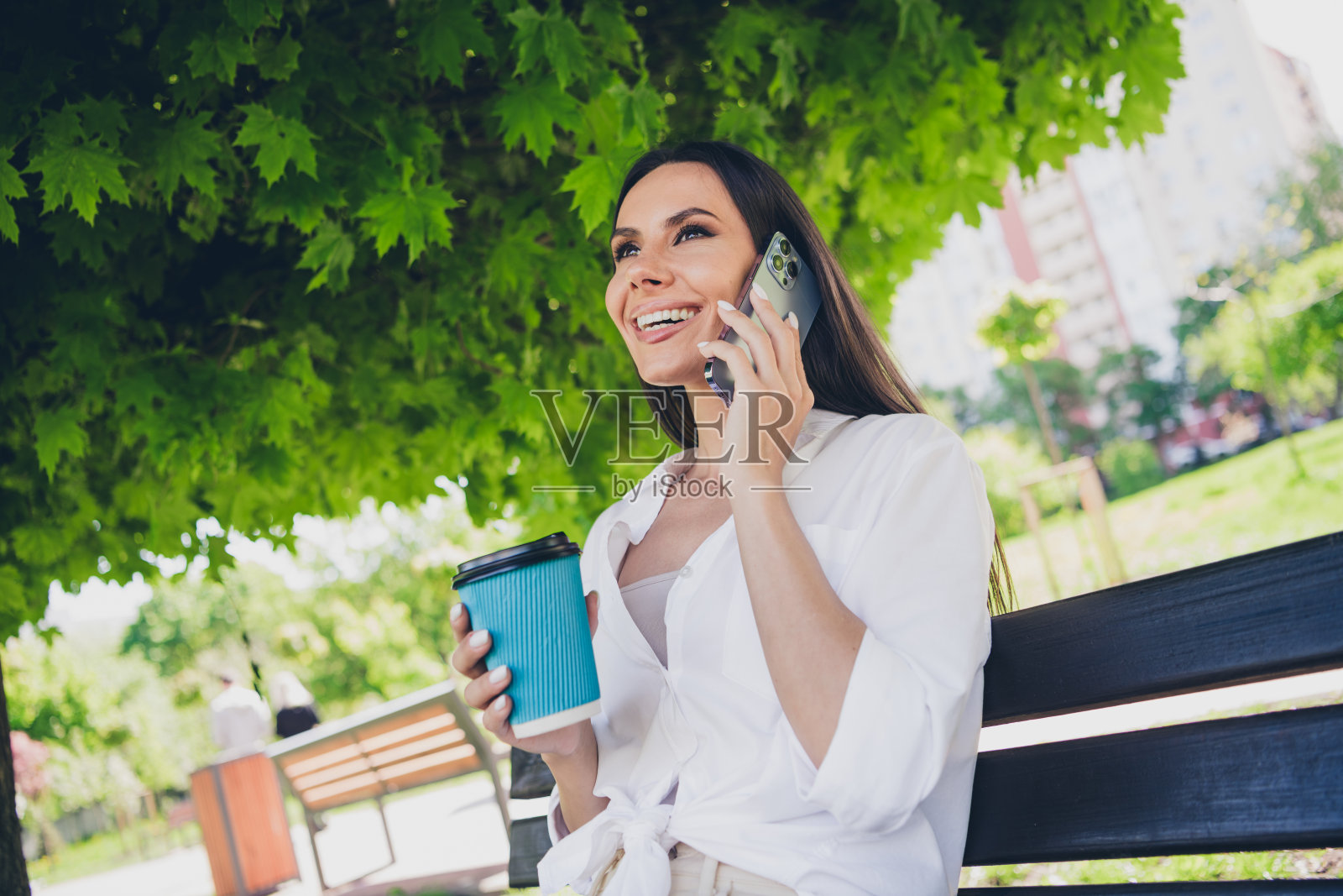 年轻快乐的女人的照片坐在长凳上讲电话喝咖啡穿着白色的衣服散步公园阳光明媚的夏天天气外面照片摄影图片