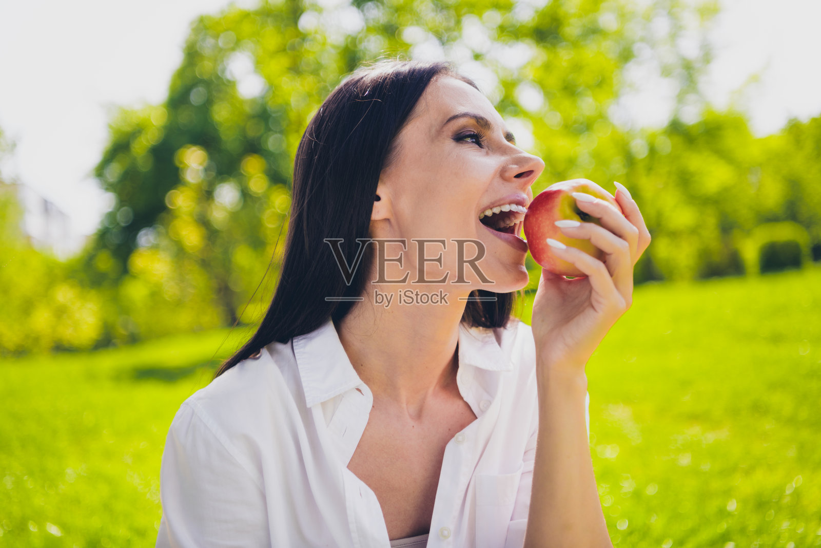 漂亮的年轻好心情的女孩吃苹果水果咬穿着白色的衣服走在城市公园外面照片摄影图片