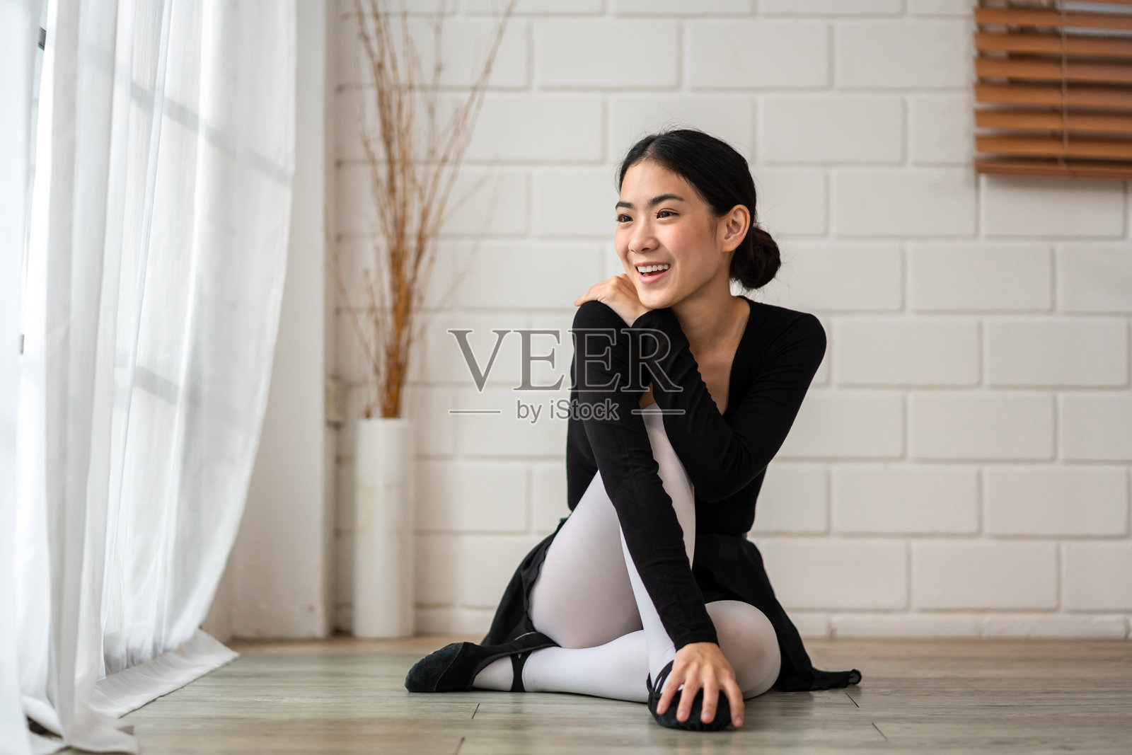 亚洲年轻芭蕾舞女演员练习芭蕾舞后坐在地板上。美丽的芭蕾舞女演员穿着芭蕾舞裙和足尖鞋，在舞厅里伸展她的脚，感到快乐和放松照片摄影图片