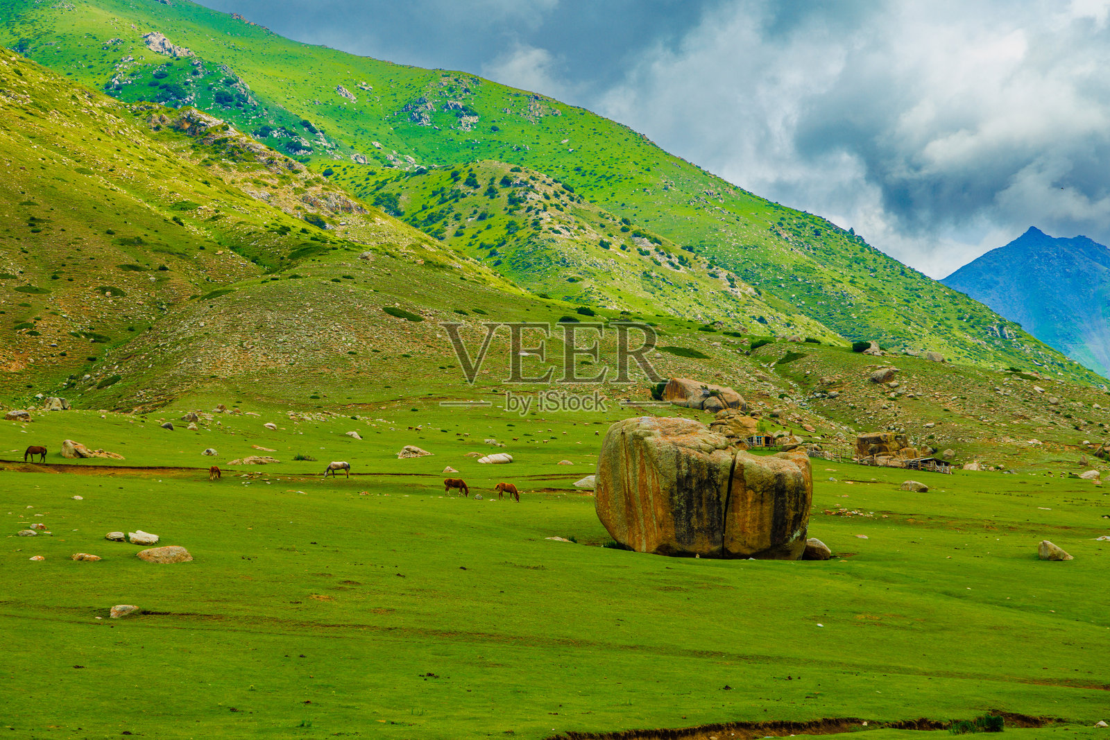 吉尔吉斯杰卢高原牧场，大岩石和几匹马在夏天的绿色山坡下吃草照片摄影图片
