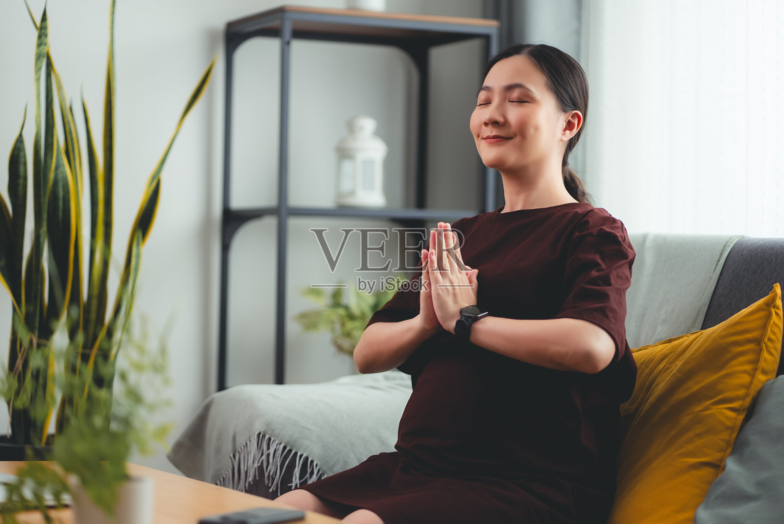 一位亚洲孕妇坐在家里的客厅里，用瑜伽的姿势握着手冥想。照片摄影图片