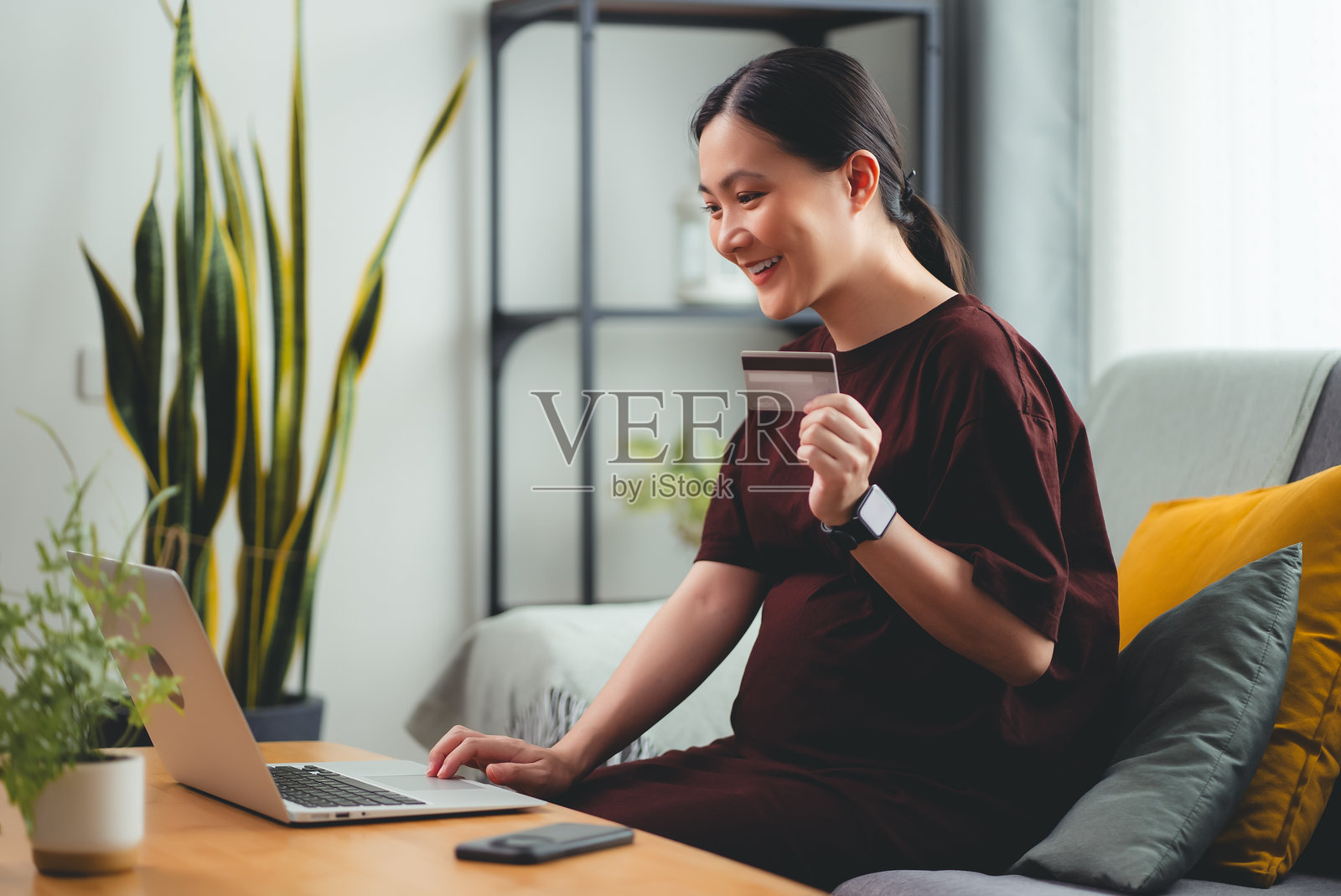 亚洲孕妇喜欢坐在家里的客厅里用笔记本电脑和信用卡进行网上购物。照片摄影图片