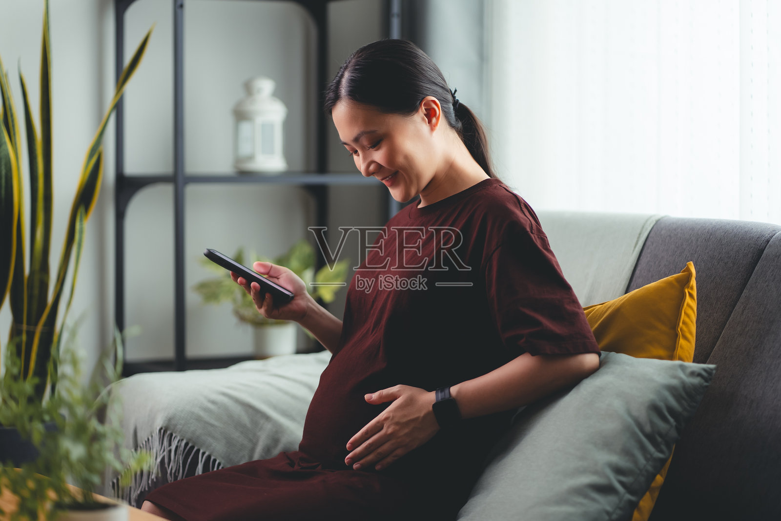 亚洲孕妇快乐地微笑着坐在沙发上，在家里使用智能手机。照片摄影图片