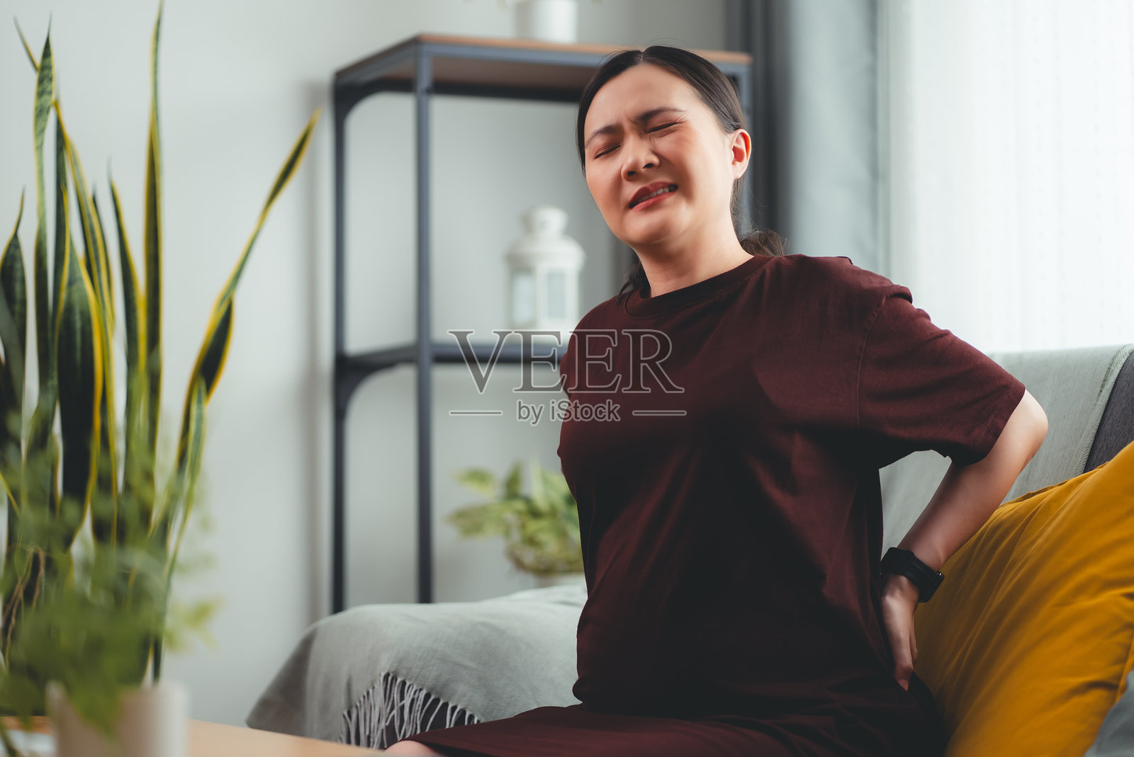 亚洲孕妇坐在家里客厅的沙发上背痛。照片摄影图片