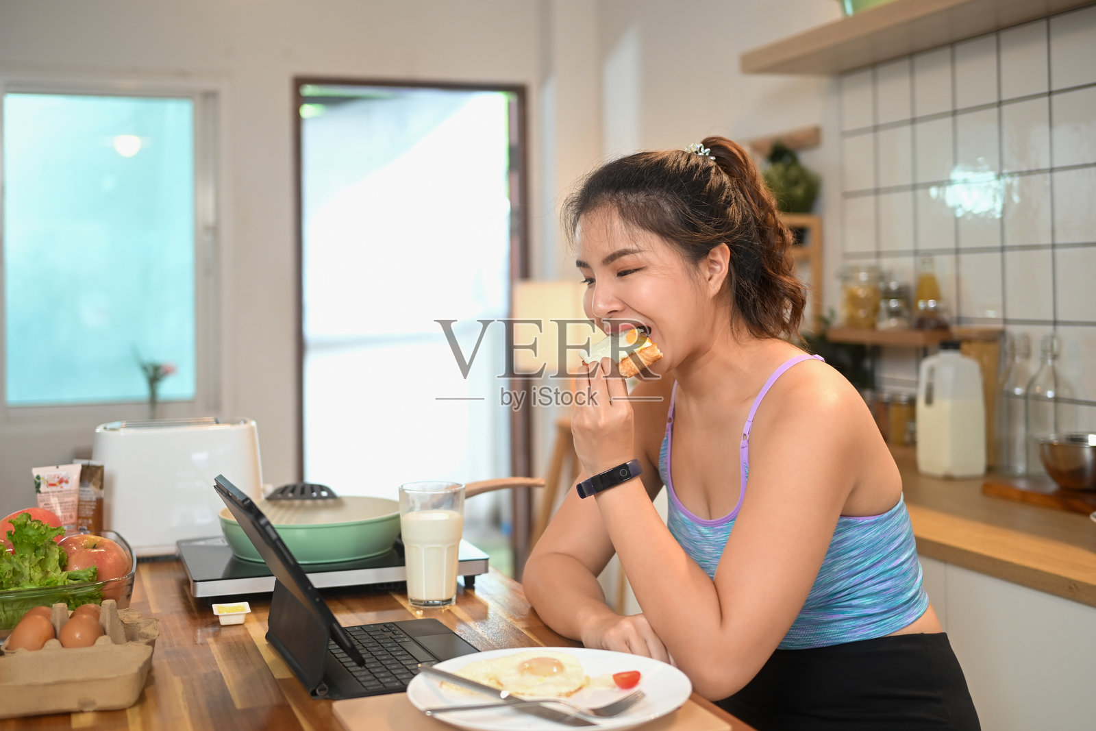 有魅力的年轻女子一边吃早餐，一边在厨房柜台上使用数码平板电脑照片摄影图片