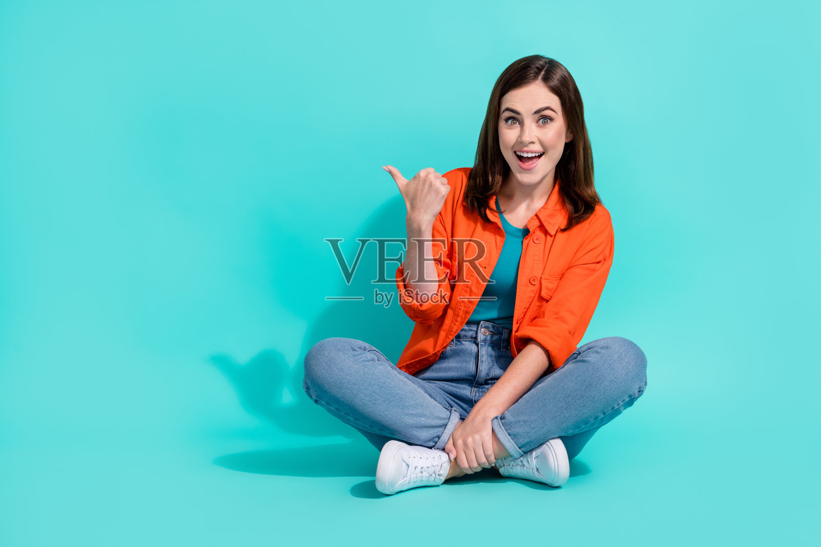 时髦的女士穿着橙色衬衫的全长照片，拇指指向空白的空间，孤立的蓝绿色背景照片摄影图片