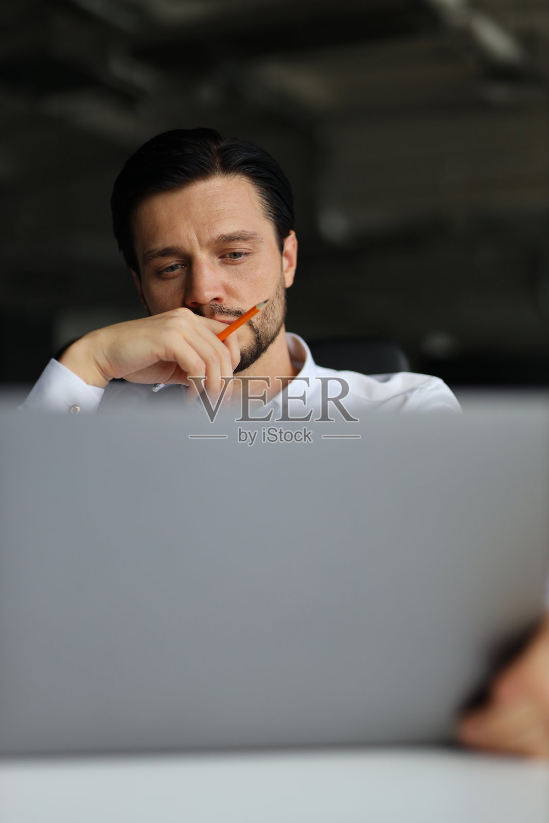 一个男人坐在办公桌前，面前放着一台打开的笔记本电脑。他嘴里叼着一支铅笔，陷入了沉思。专注和沉思的概念照片摄影图片