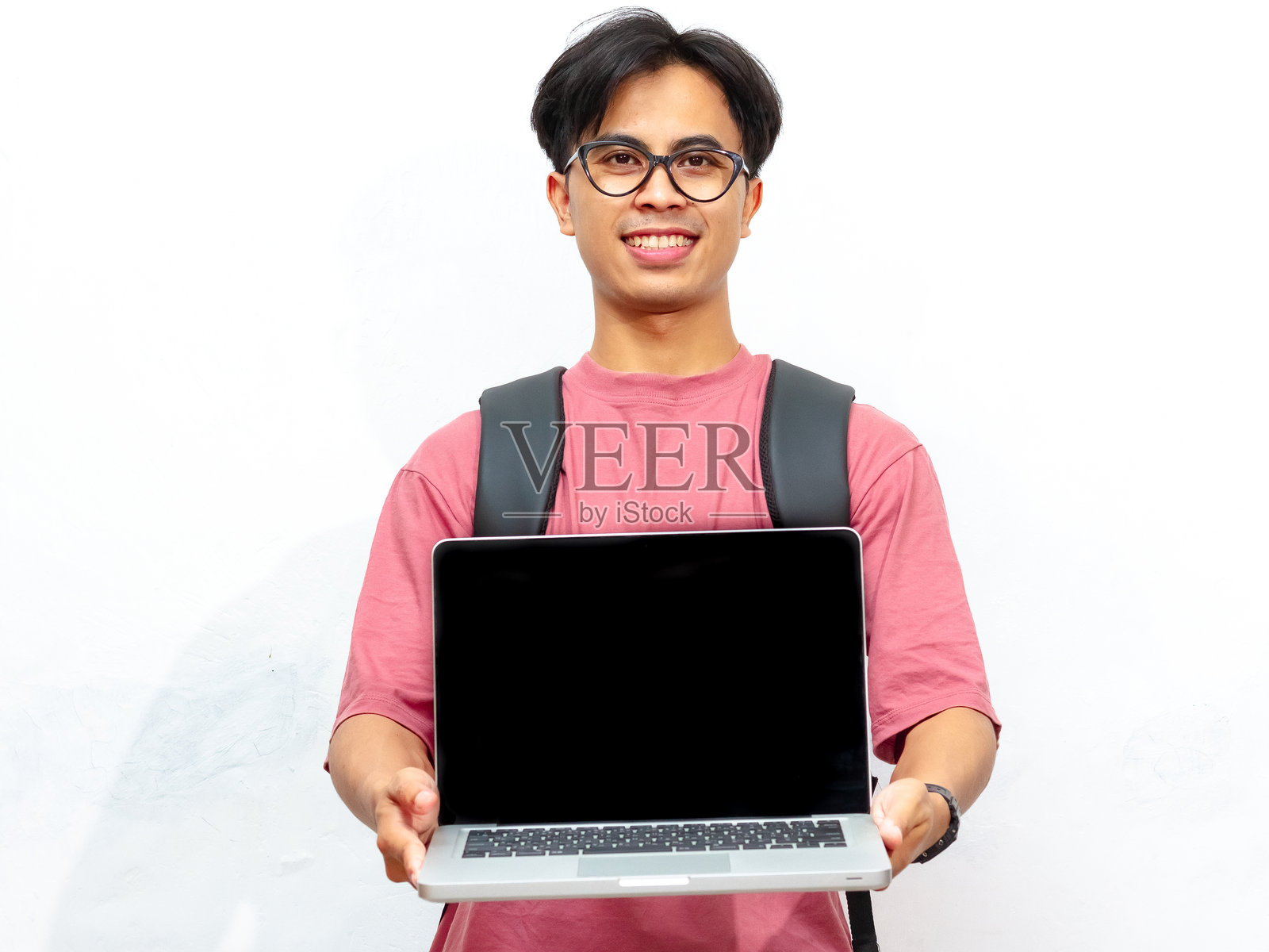 一个年轻的亚洲男学生的肖像，穿着粉红色的t恤，背着背包，兴奋地拿着一台笔记本电脑，白底上有一个空白的屏幕。照片摄影图片