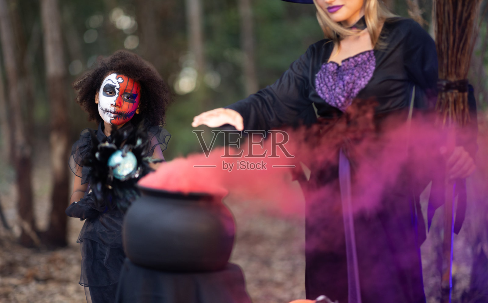 穿着女巫服装的无忧无虑的母亲和化妆成骷髅头的女儿，在森林里用沸腾的锅，喷出五颜六色的烟雾，尽情地表演魔术照片摄影图片