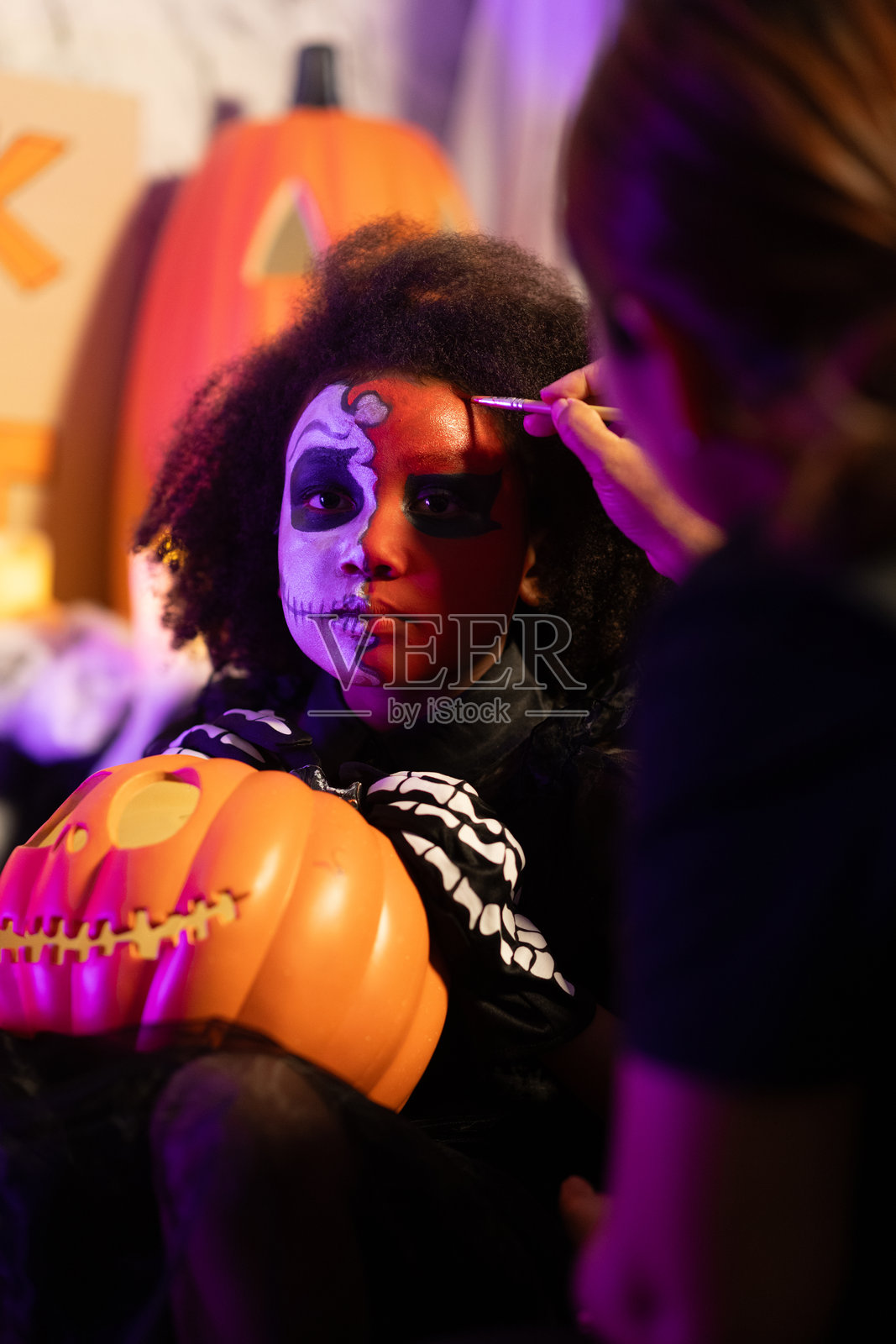 万圣节派对。为了庆祝亡灵节，化妆师正在给一个黑人女孩画骷髅。一个化着骷髅妆的黑人女孩。万圣节化妆和服装概念。照片摄影图片