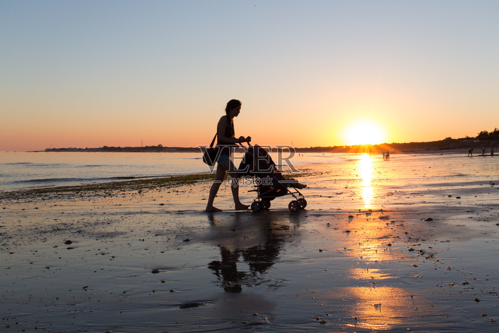 在日落期间，推着婴儿婴儿车的妇女在海滩上的晴空照片摄影图片
