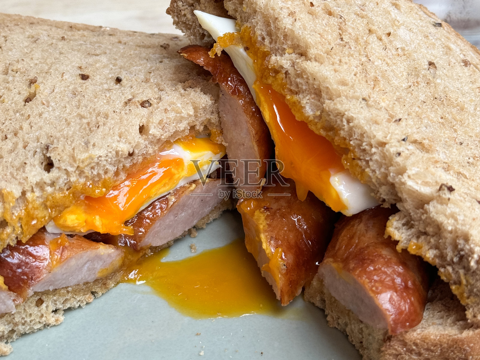 全景图：全麦面包香肠和煎蛋三明治放在白色盘子上，三角形的三明治切成两半，蛋黄流淌，焦点在前景。照片摄影图片