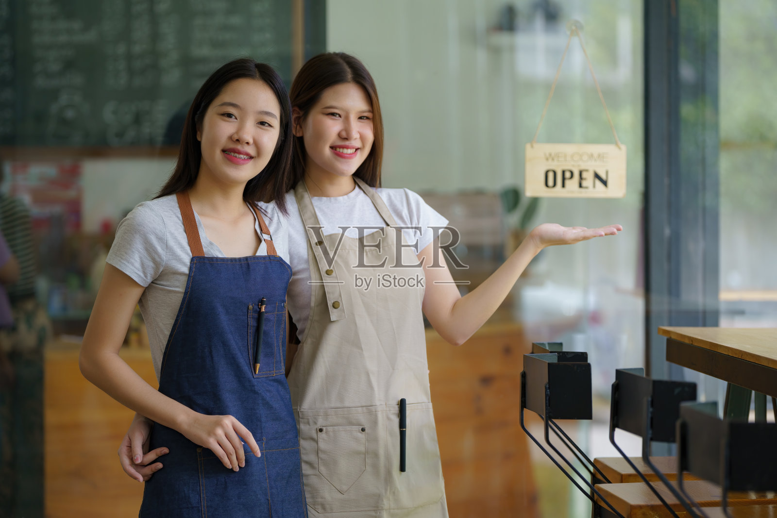 两位亚洲女咖啡师自信地站在咖啡店前，微笑着看向镜头，阳光明媚。照片摄影图片