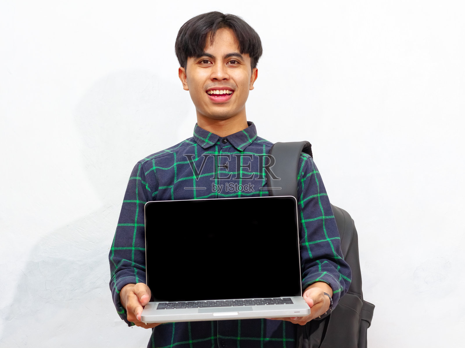微笑自信的亚洲男性学生手持背包，展示空白的笔记本电脑屏幕，背景为白色。非常适合广告使用。照片摄影图片