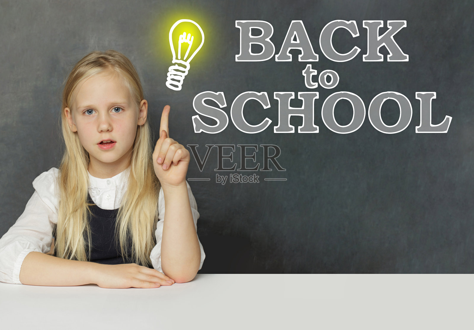 快乐的小学生女孩用手指指着黑板背景上的黄色灯泡，黑板上写着“开学季”的字样。教育概念。照片摄影图片