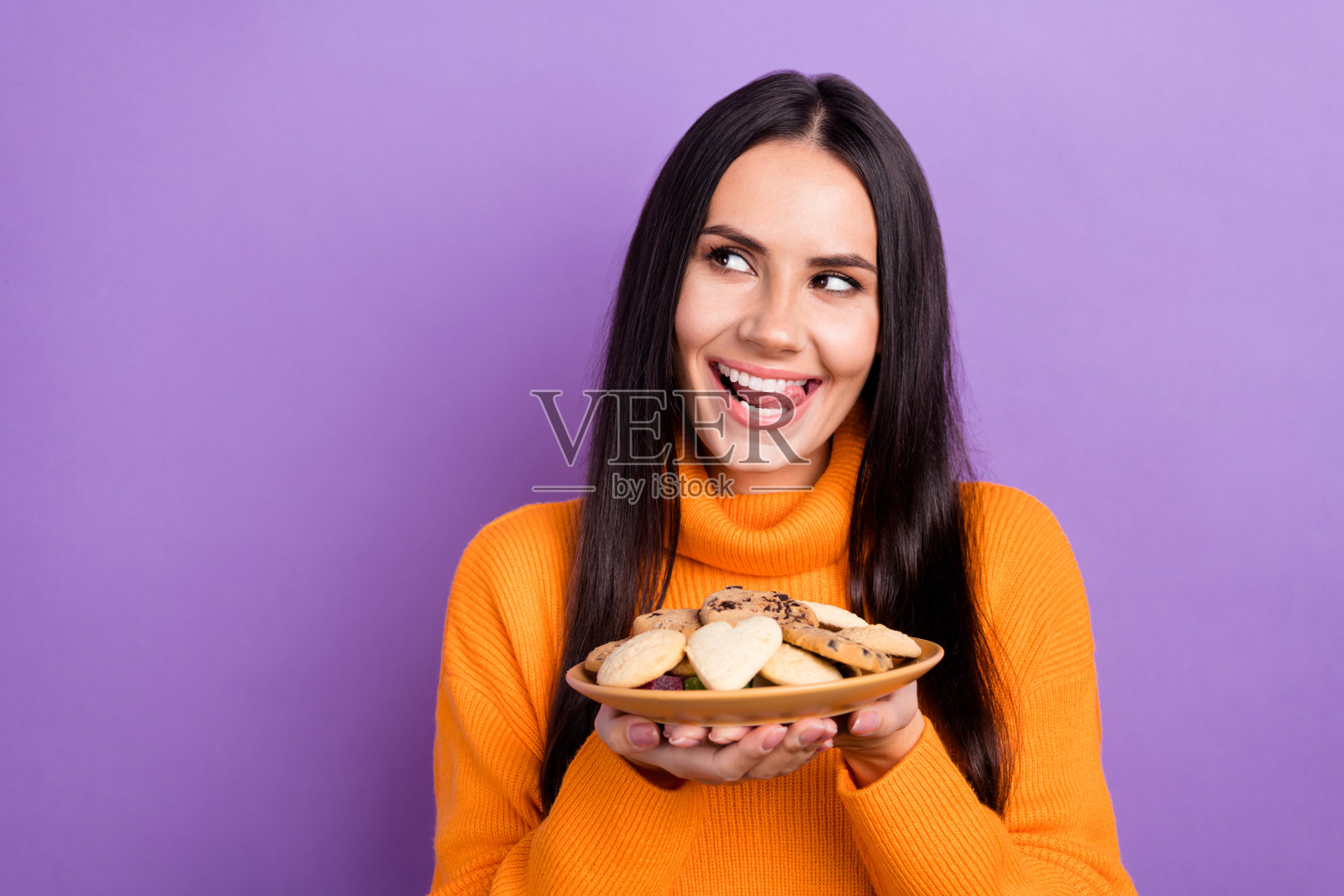 饥饿的棕发女人穿着时髦的橙色毛衣，伸出舌头，拿着饼干，紫色背景照片摄影图片