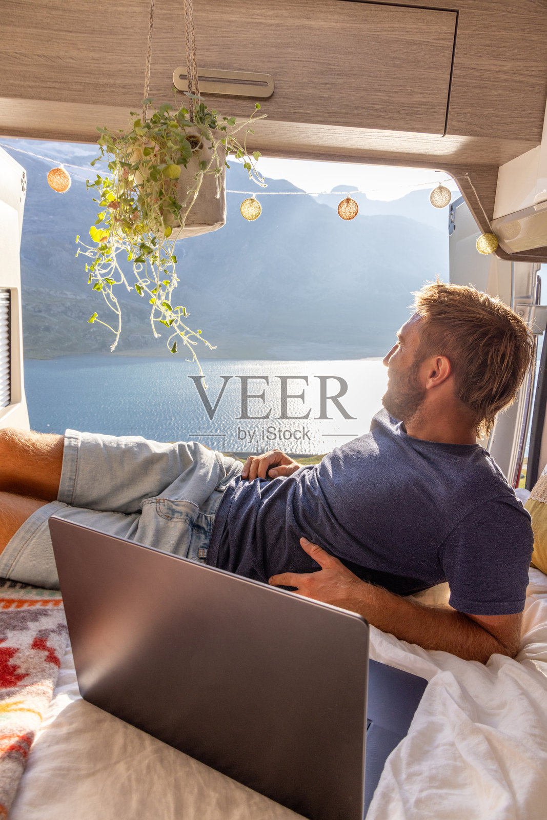 男子在露营车床上放松，手里拿着笔记本电脑，窗外是风景如画的湖泊景色照片摄影图片