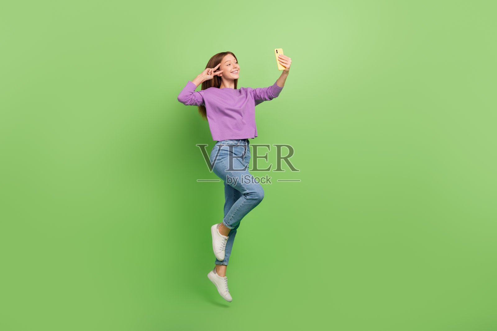 全身照，穿着牛仔裤和紫色毛衣的可爱积极女孩，手持智能手机做出V字手势，背景为绿色。照片摄影图片